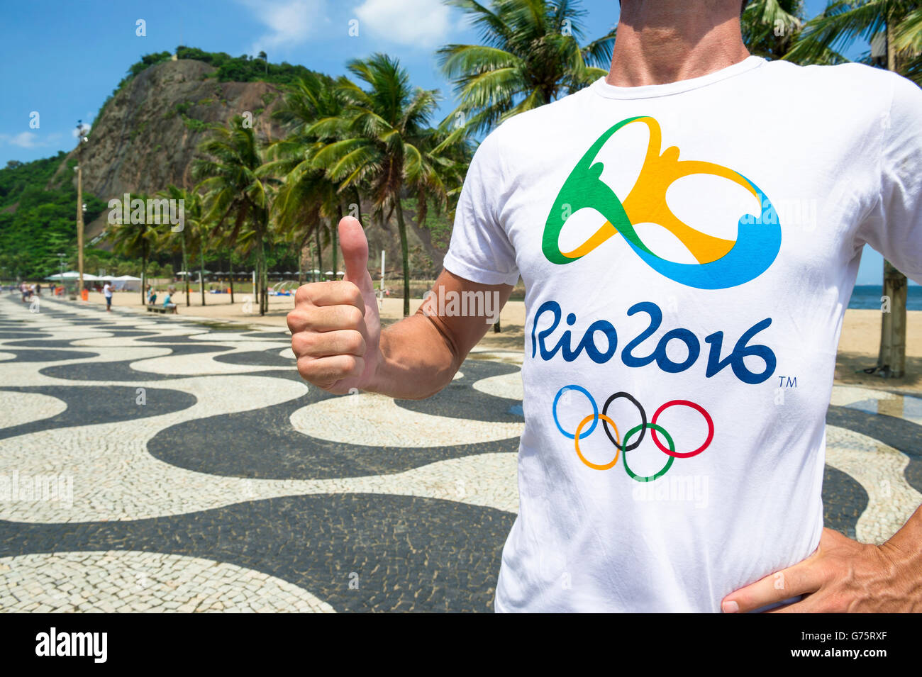 RIO DE JANEIRO - 10. März 2016: Mann mit offiziellen steht Rio 2016 T-shirt einen Daumen nach Copacabana Strand Gehweg aufzugeben. Stockfoto