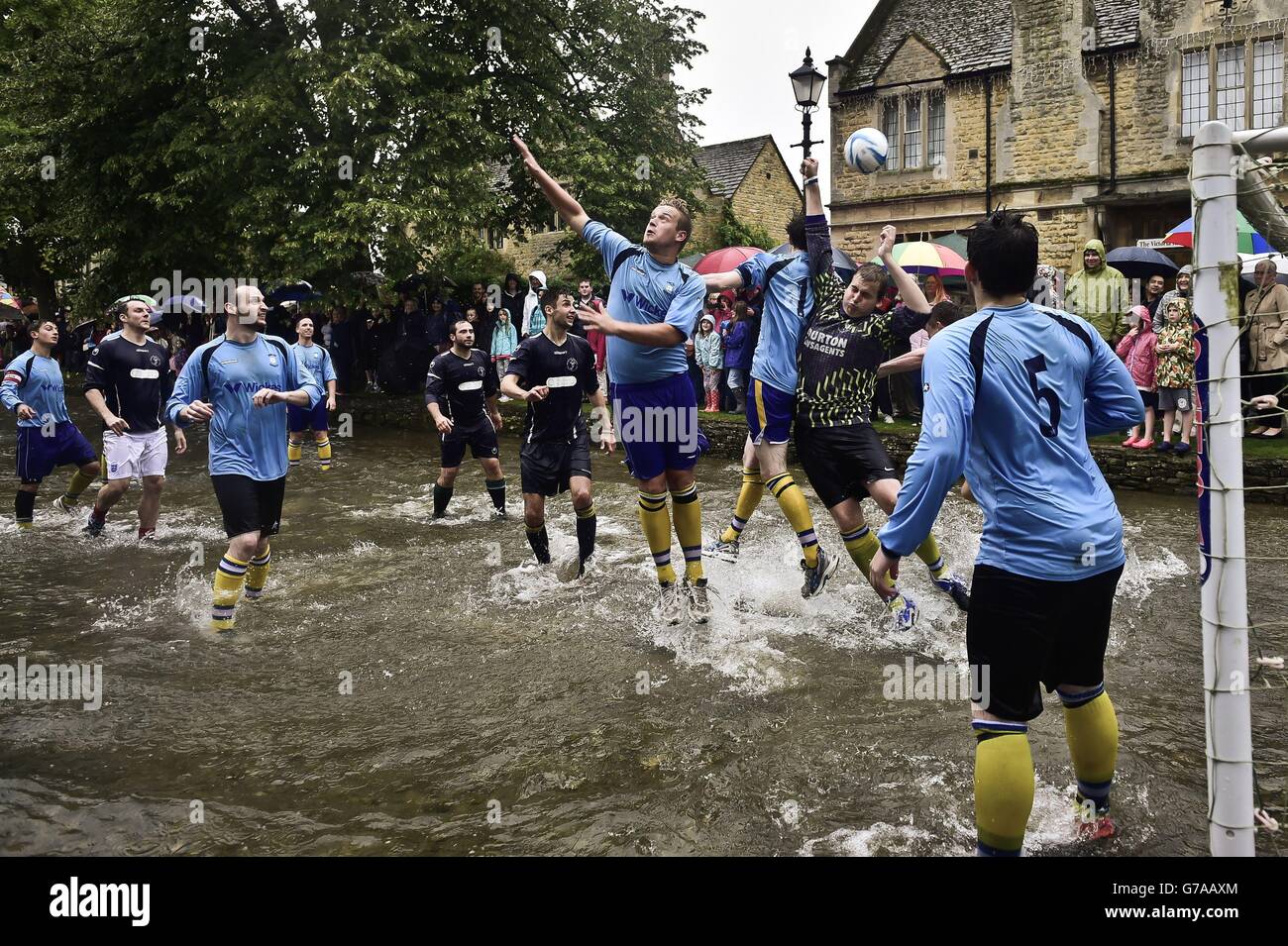 Der Ball kommt über die Spitze, als Bourton Rovers 1st XI (dunkelblau) und Bourton Rovers 2nd XI (hellblau) beim jährlichen Football in the River-Spiel in Bourton-in-the-Water, Gloucestershire, Wo im River Windrush auf der Hauptstraße im Dorf zwei 15-minütige Fußballhälften gespielt werden. Stockfoto