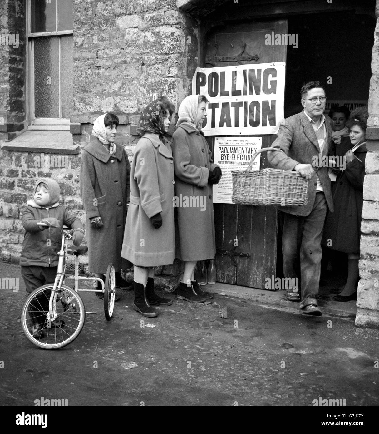 Ein Bäcker mit einem Korb am Arm verlässt ein Wahllokal in Towcester, nachdem er in der Nachwahl in South Northamptonshire gewählt hat. Frauen in Kopftüchern stehen vor der Tür, während ein kleines Kind und zukünftiger Wähler sein Dreirad anhält und zuschaut. Stockfoto
