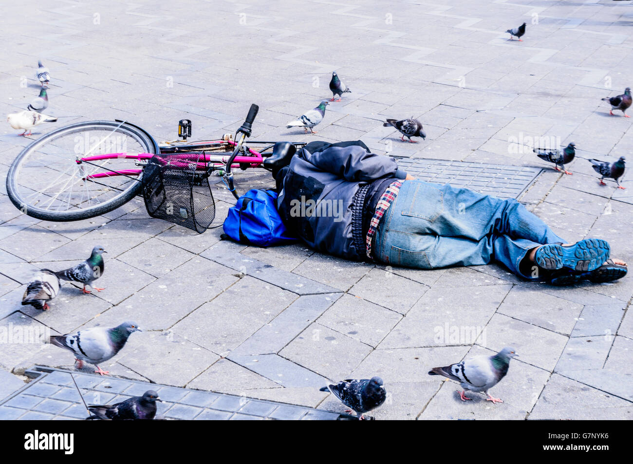 Ein betrunkener Mann schläft auf einem öffentlichen Platz neben seinem Fahrrad, während Tauben um ihn herum laufen. Stockfoto