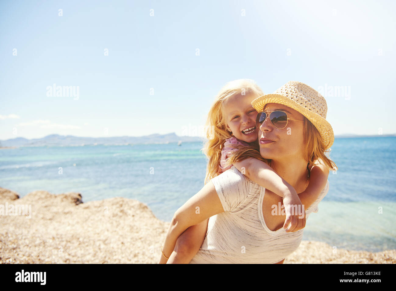 Lachend temperamentvoll wenig blondes Mädchen mit ihrer Mutter am Strand Reiten auf ihr im Huckepack auf einem felsigen Ufer, Ozean Hintergrund Stockfoto