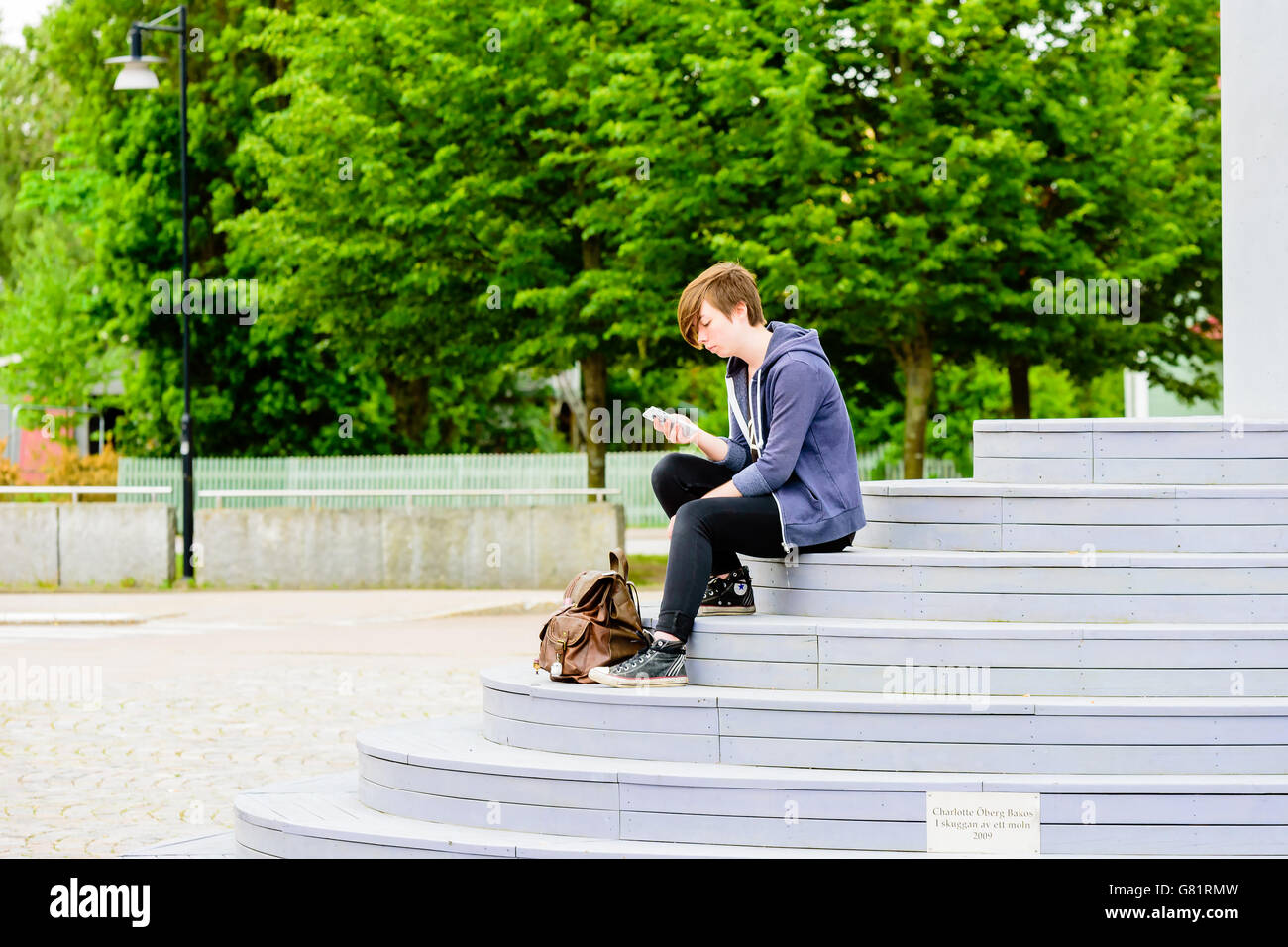 Söderköping, Schweden - 20. Juni 2016: Junge Erwachsene Person sitzt auf der Treppe, ein künstlerisches Denkmal während SMS auf das mobil Stockfoto