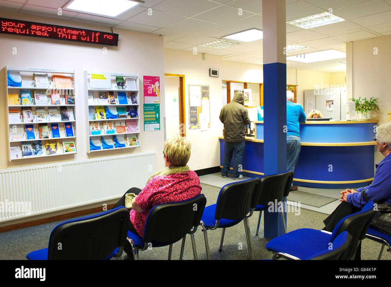 Wartezimmer in einer britischen GP-Praxis mit Patienten sitzen und stehen durch die Rezeption, die darauf warten, registrieren Stockfoto