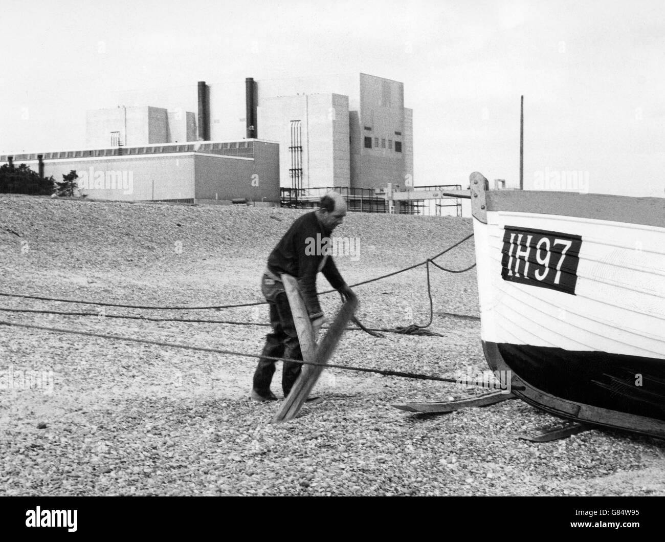 Ein Suffolk-Fischer beobachtet, wie sein Boot The Four Sisters mit einem Scholle-Fang an Bord den Kiesstrand von Sizewell, Suffolk, hinauffährt. Im Hintergrund sind die Gebäude des Kernkraftwerks Sizewell zu sehen, das im vergangenen Monat offiziell eröffnet wurde. Stockfoto