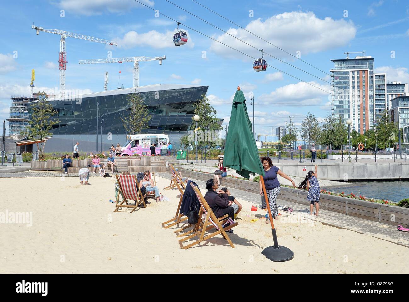 Familien entspannen sich auf den kostenlosen Liegestühlen und genießen die Sonne am künstlichen Strand am westlichen Ende der Royal Victoria Docks, in Newham, im Osten Londons, unterhalb der Emirates Air Line. Während des letzten heißen Wetters wurde eine Warteschlange von 1,000 Personen aufgezeichnet. Stockfoto