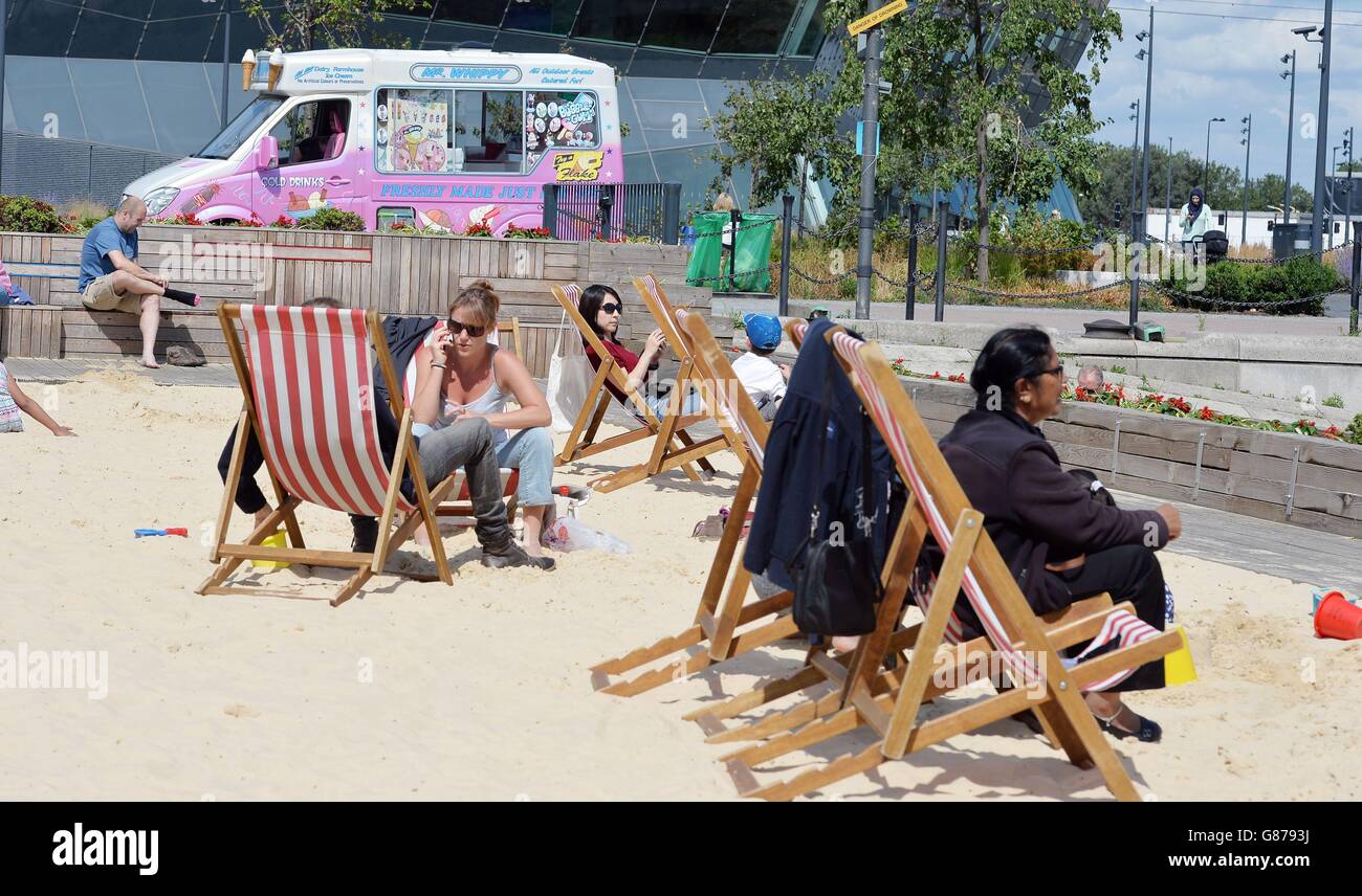 Familien entspannen sich auf den kostenlosen Liegestühlen und genießen die Sonne am künstlichen Strand am westlichen Ende der Royal Victoria Docks in Newham, Ost-London, wo bei kürzlichem heißem Wetter eine Schlange von 1,000 Personen aufgezeichnet wurde. Stockfoto