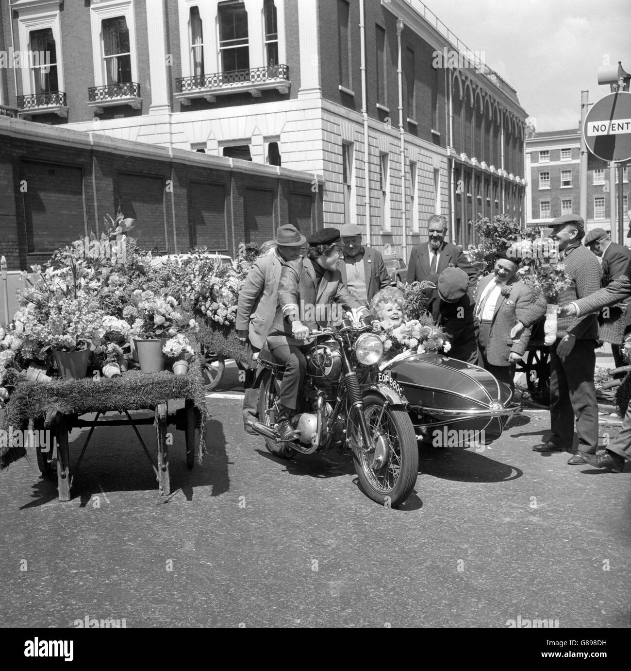 Lucille Ball fährt im Beiwagen des Motorrads, das von dem britischen Anthony Newley während der Dreharbeiten am Manchester Square für eine Fernsehserie „Lucy in London“ gefahren wird. Stockfoto