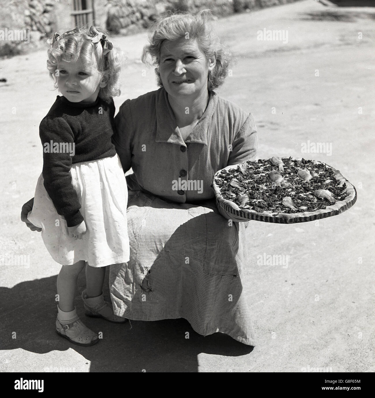 der 1950er Jahre, historisch, mallorquinischen Dame mit Kind zeigt ein traditionelles lokales Gericht, ein Coca de Trampo, ähnlich wie eine Pizza, aber ohne Käse und mehr Gemüse. Stockfoto