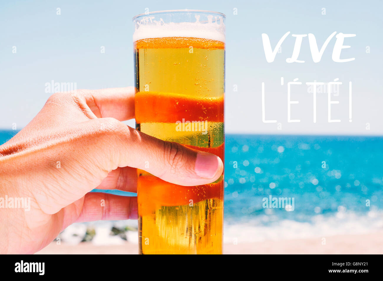 Nahaufnahme eines kaukasischen Jünglings mit einem Glas mit erfrischendem Bier in der Hand mit dem Meer im Hintergrund und der Text viv Stockfoto
