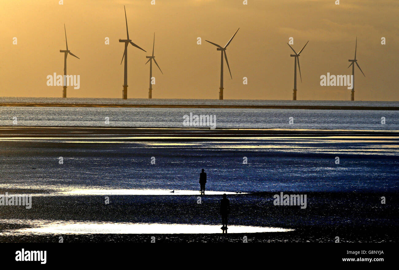 Die Sonne geht über Windturbinen unter, die man vom Strand in Crosby, Merseyside, aus sehen kann, während zwei von Antony Gormleys Iron Men auf das Meer blicken. Stockfoto