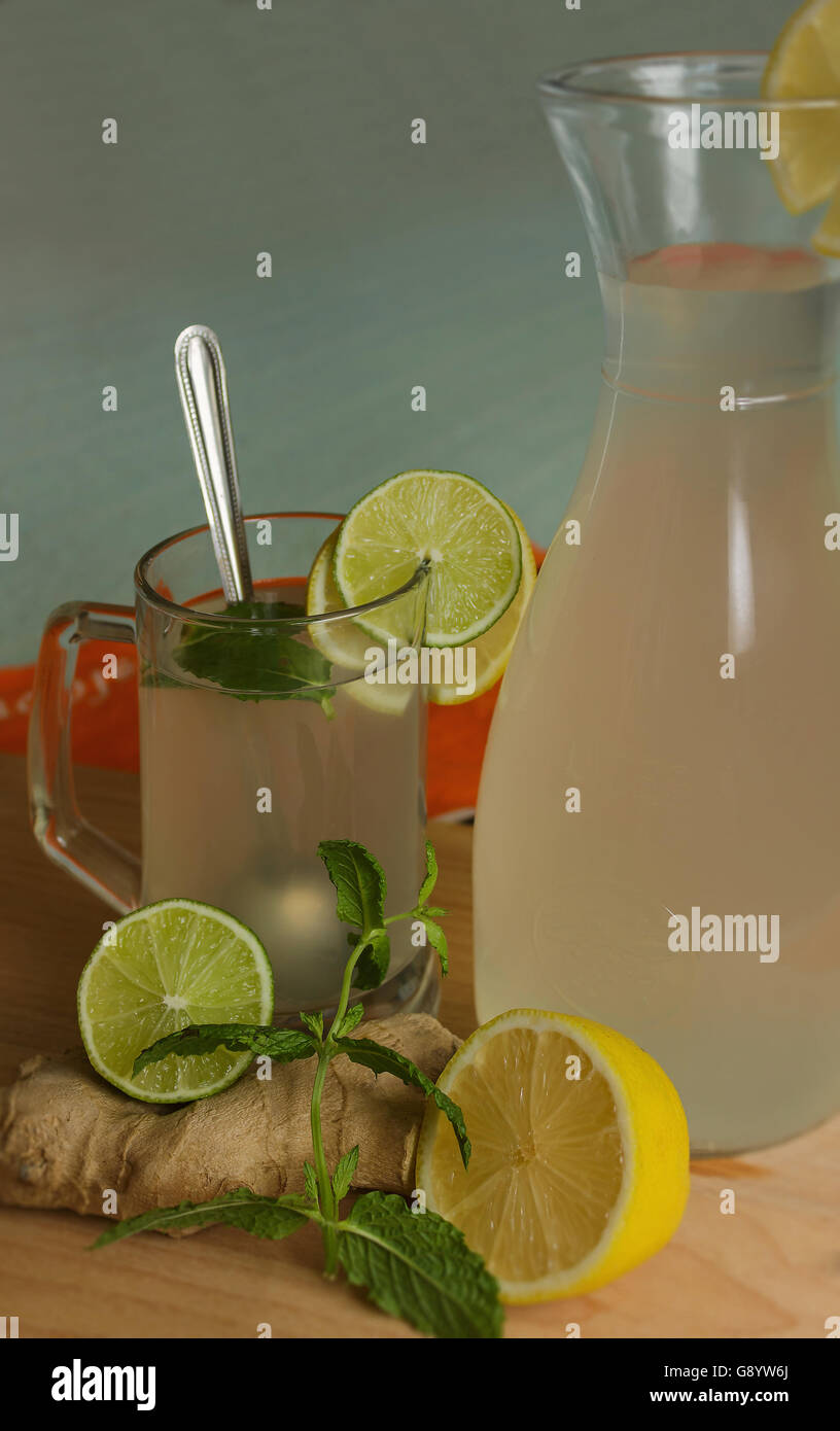 Ingwer und Zitrone Saft in ein Glas und Krug Plank Ingwer, Zitrone, Limette und Minze Blätter auf einem farbigen Hintergrund vertikale Ansicht Stockfoto