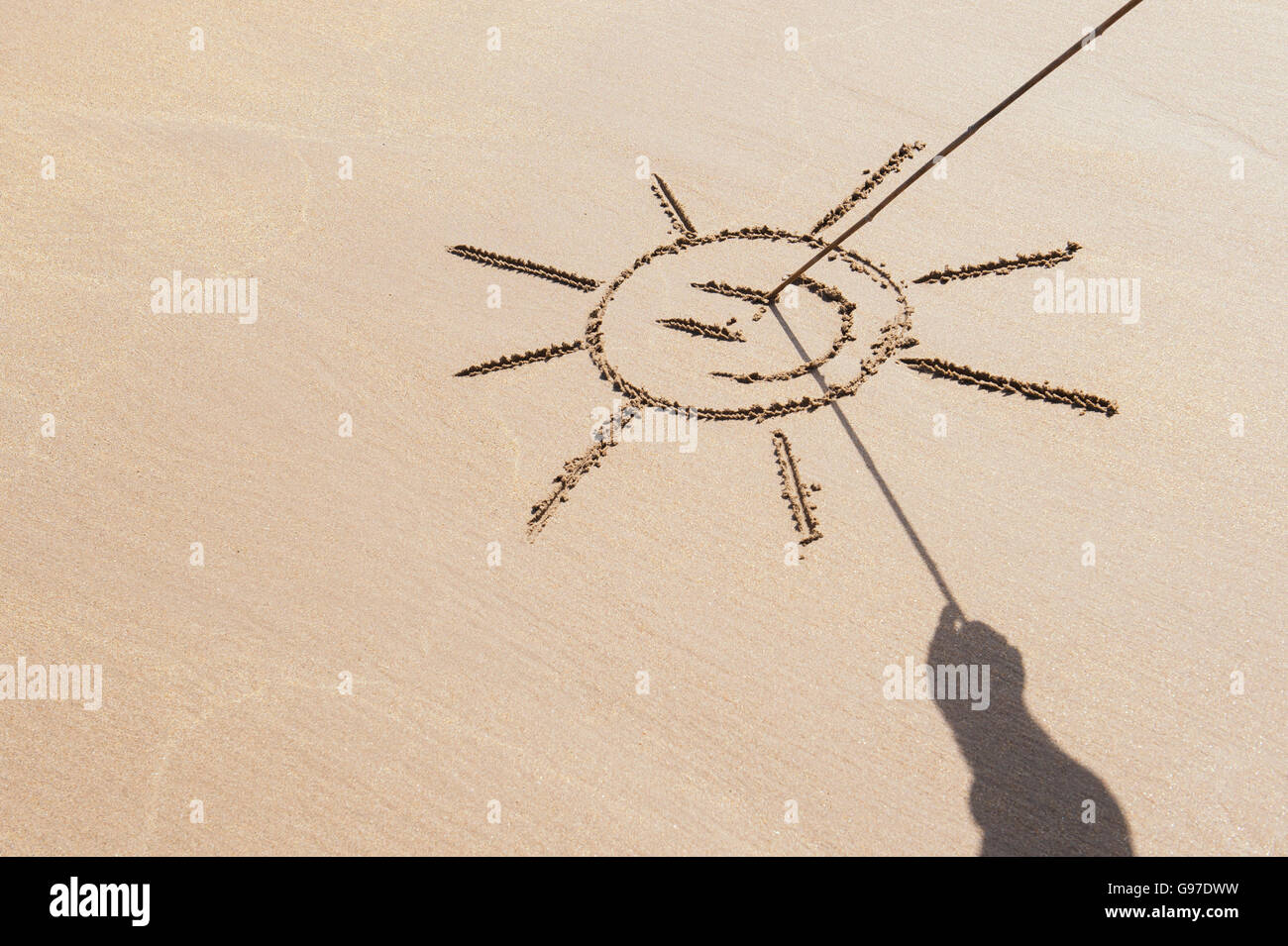 Schatten-Hand hält einen Stock zeichnen eine Smiley-Gesicht-Sonne am Strand. UK Stockfoto