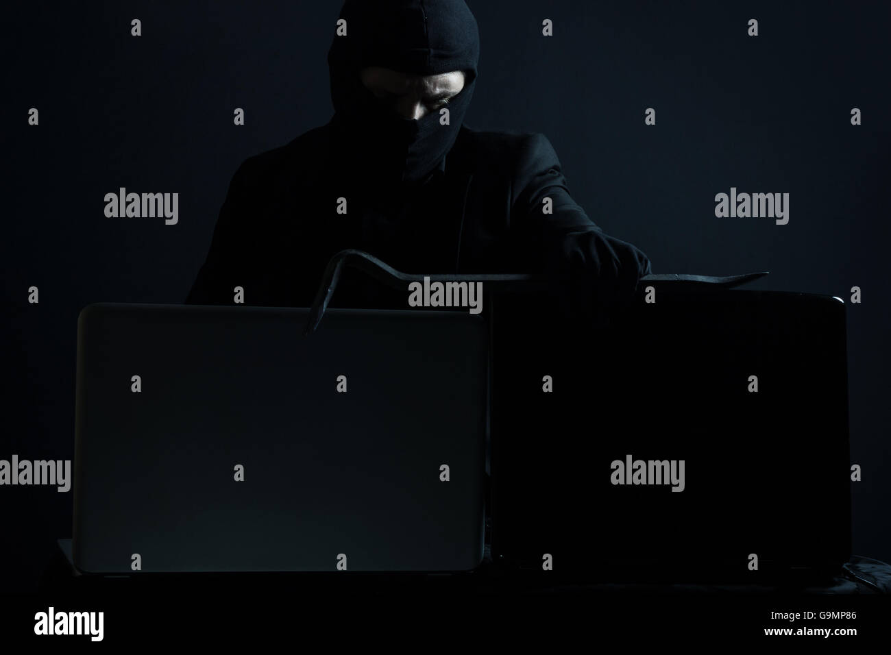 Böse Computerhacker in Anzug stehlen Daten von Laptop mit Brecheisen vor schwarzem Hintergrund Stockfoto