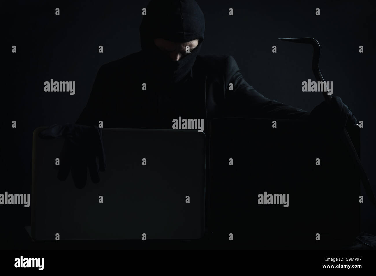 Böse Computerhacker in Anzug stehlen Daten von Laptop mit Brecheisen und Handschuhe vor schwarzem Hintergrund Stockfoto
