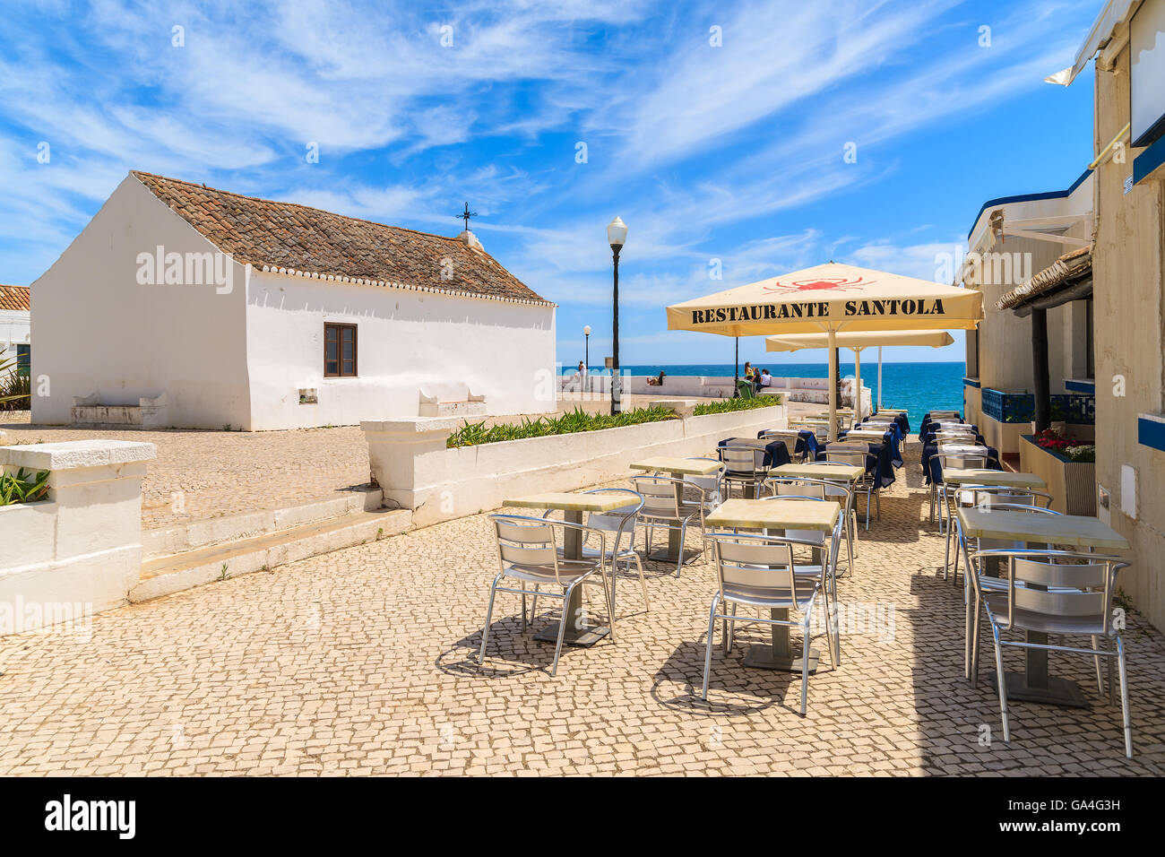 ARMACAO DE PERA Stadt, PORTUGAL - 17. Mai 2015: Restauranttische und weiße Kirche auf Küstenpromenade in Armacao de Pera Küstenstadt. Die Region Algarve ist beliebtes Urlaubsziel für Touristen im Sommer. Stockfoto