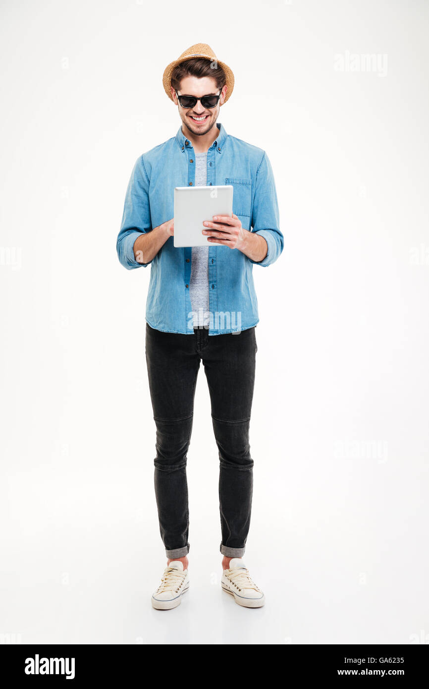 Freundlichen jungen Mann in Hut und eine Sonnenbrille stehend und mit Tablet auf weißem Hintergrund Stockfoto