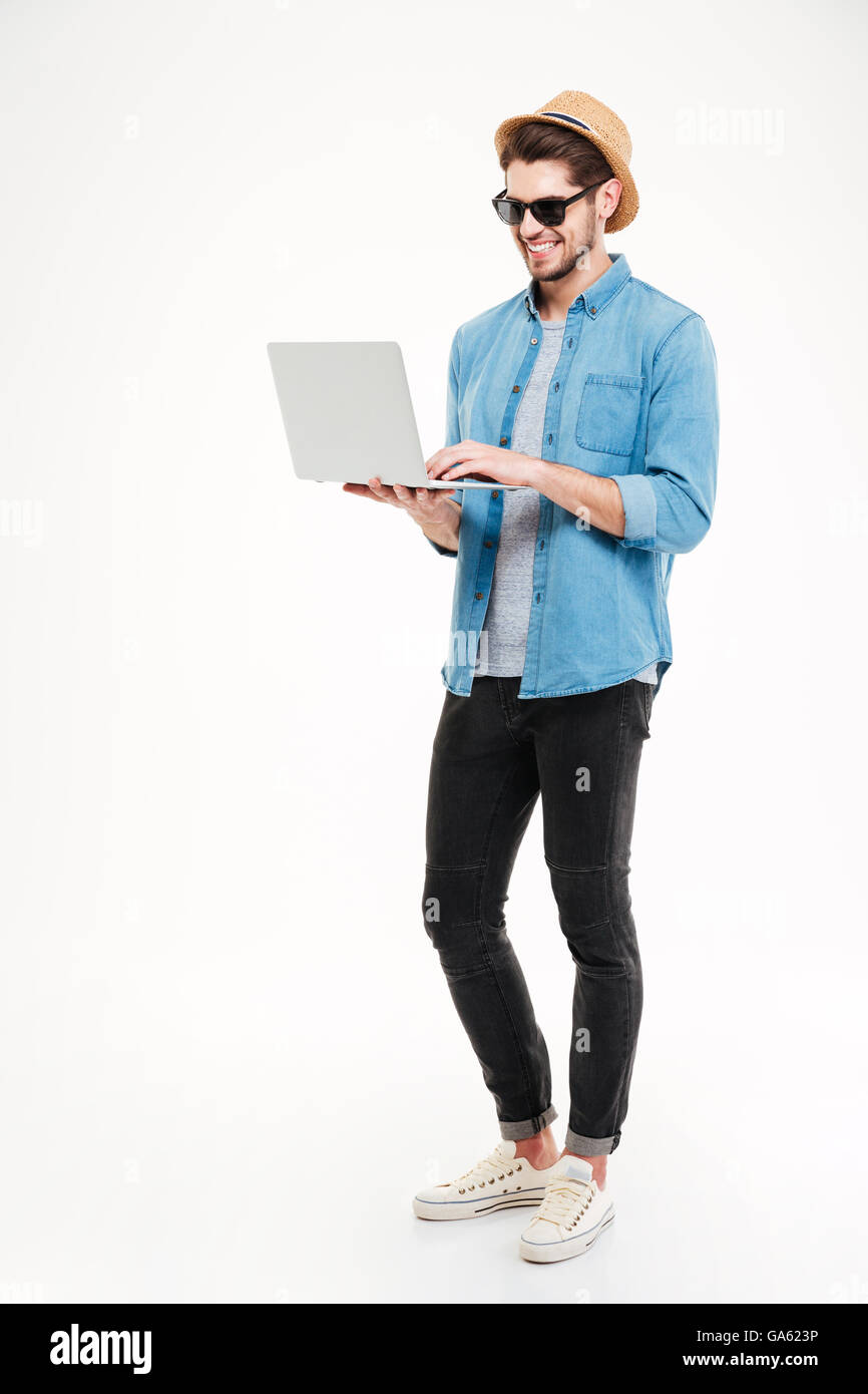 Fröhlichen jungen Mann in Hut und eine Sonnenbrille stehend und mit Laptop auf weißem Hintergrund Stockfoto