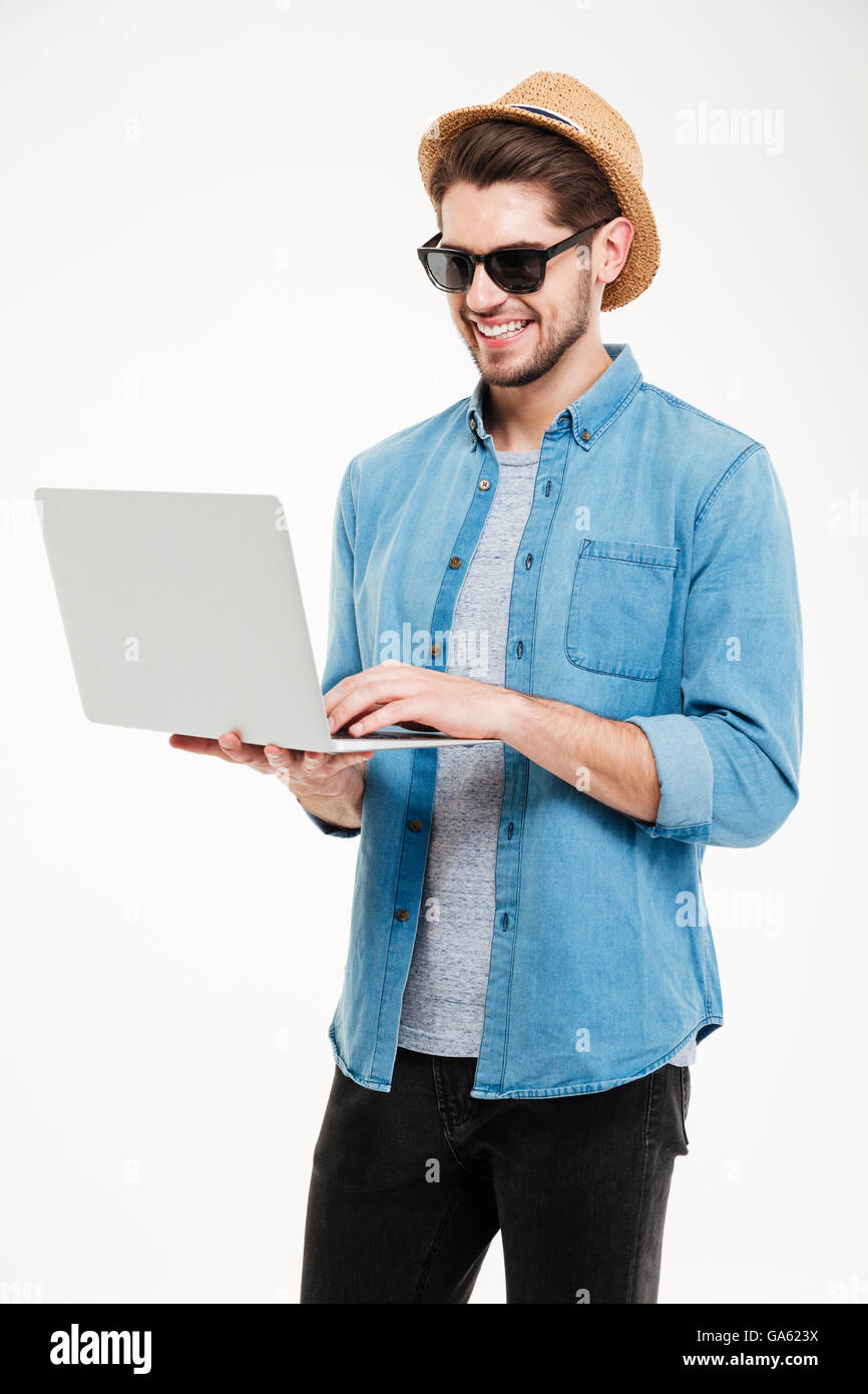 Lächelnde attraktiver junger Mann mit Sonnenbrille und Hut stehend und mit laptop Stockfoto