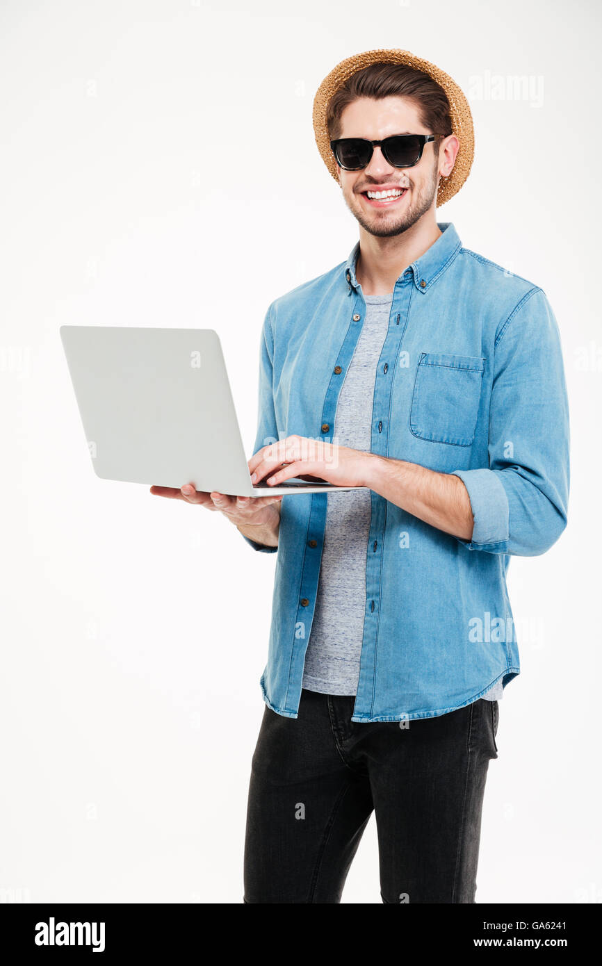 Glücklich hübscher junger Mann mit Sonnenbrille und Hut hält und mit laptop Stockfoto
