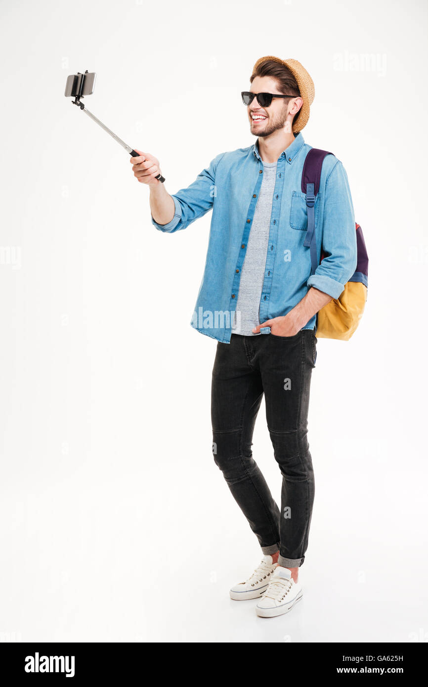 Voller Länge lächelnder jungen Mann mit Rucksack fotografieren mit Smartphone und Selfie stick Stockfoto