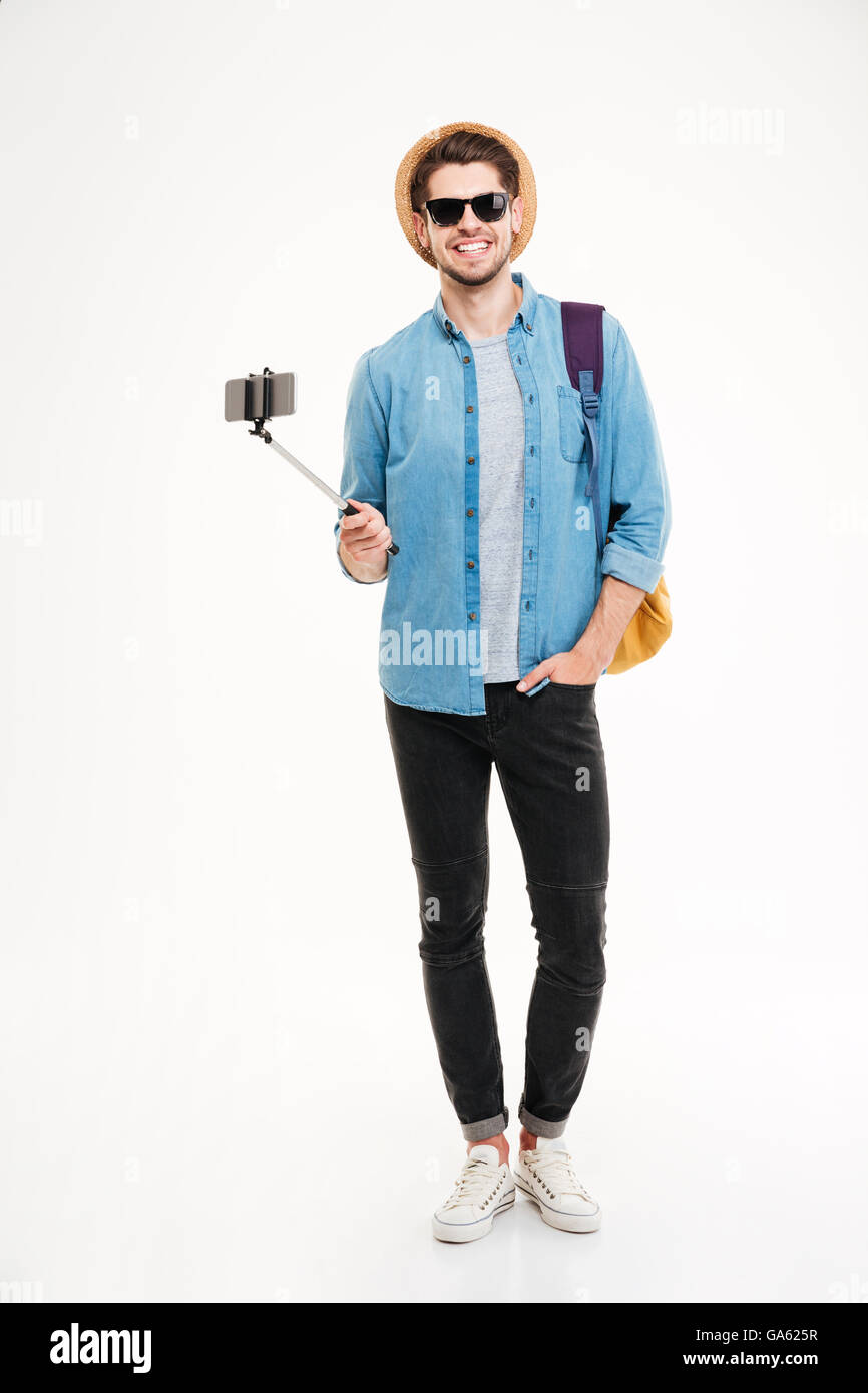 Voller Länge lächelnder jungen Mann mit Rucksack und Handy auf Selfie Stick auf weißem Hintergrund Stockfoto