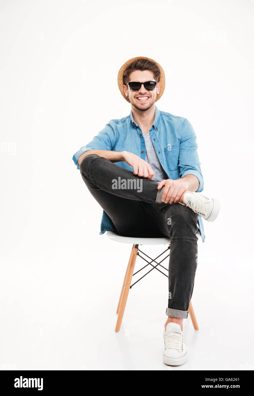 Fröhliche entspannte junger Mann in Hut und Brille auf Stuhl sitzend, auf weißem Hintergrund Stockfoto