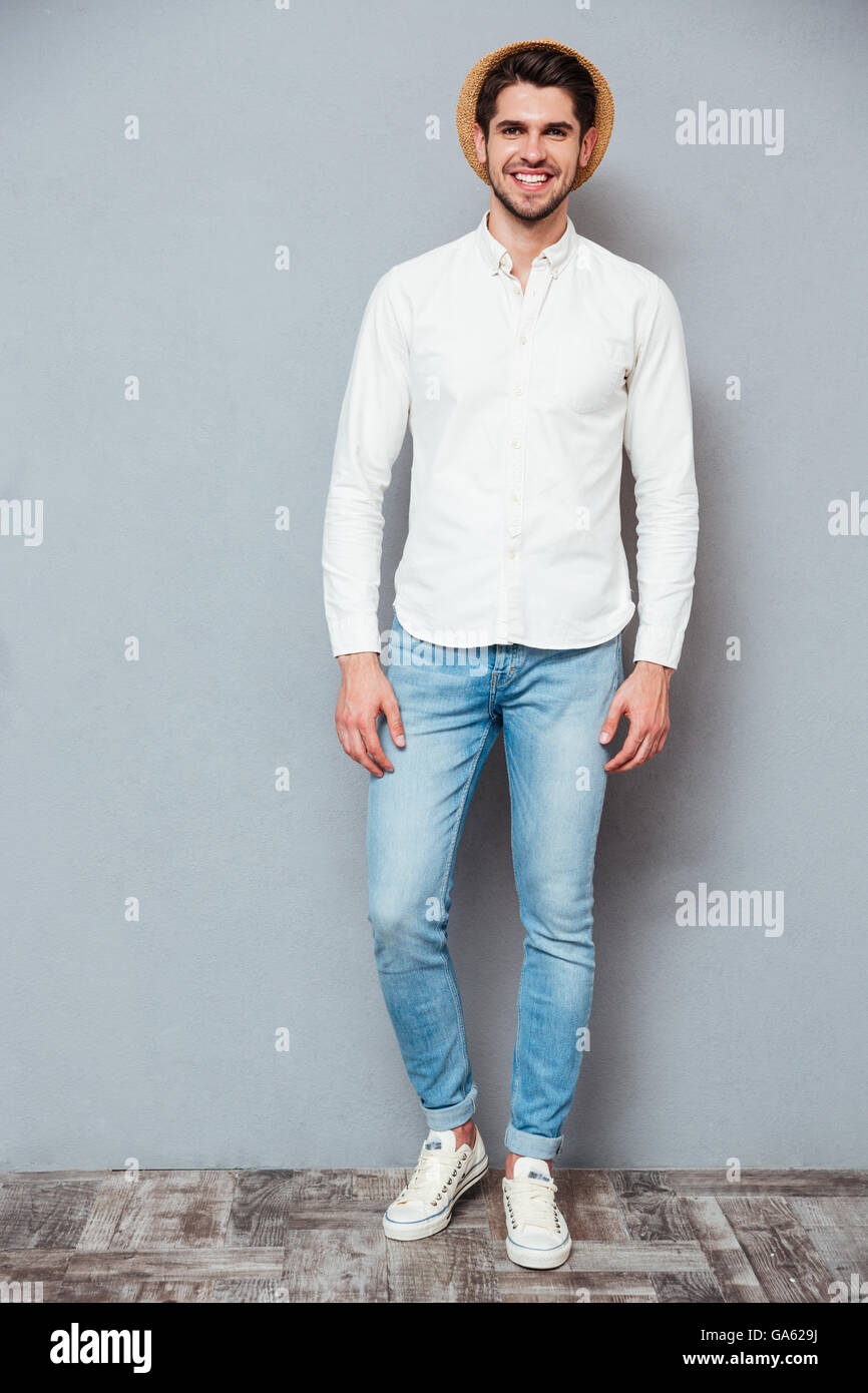 Voller Länge Porträt des Lächelns hübschen jungen Mannes im weißen Hemd, Jeans und Hut stehen auf grauem Hintergrund Stockfoto