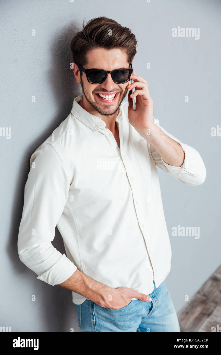 Fröhlich attraktive junge Mann im weißen Hemd und Sonnenbrille telefonieren mit Handy über grauen Hintergrund Stockfoto