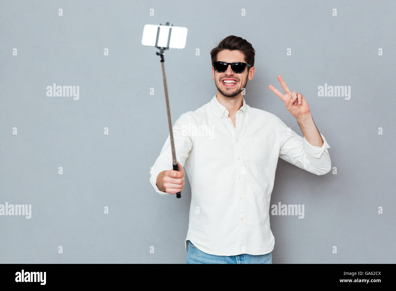 Lächelnd jungen Mann mit Sonnenbrille fotografieren mit Smartphone und Selfie Stick auf einem grauen Hintergrund isoliert Stockfoto