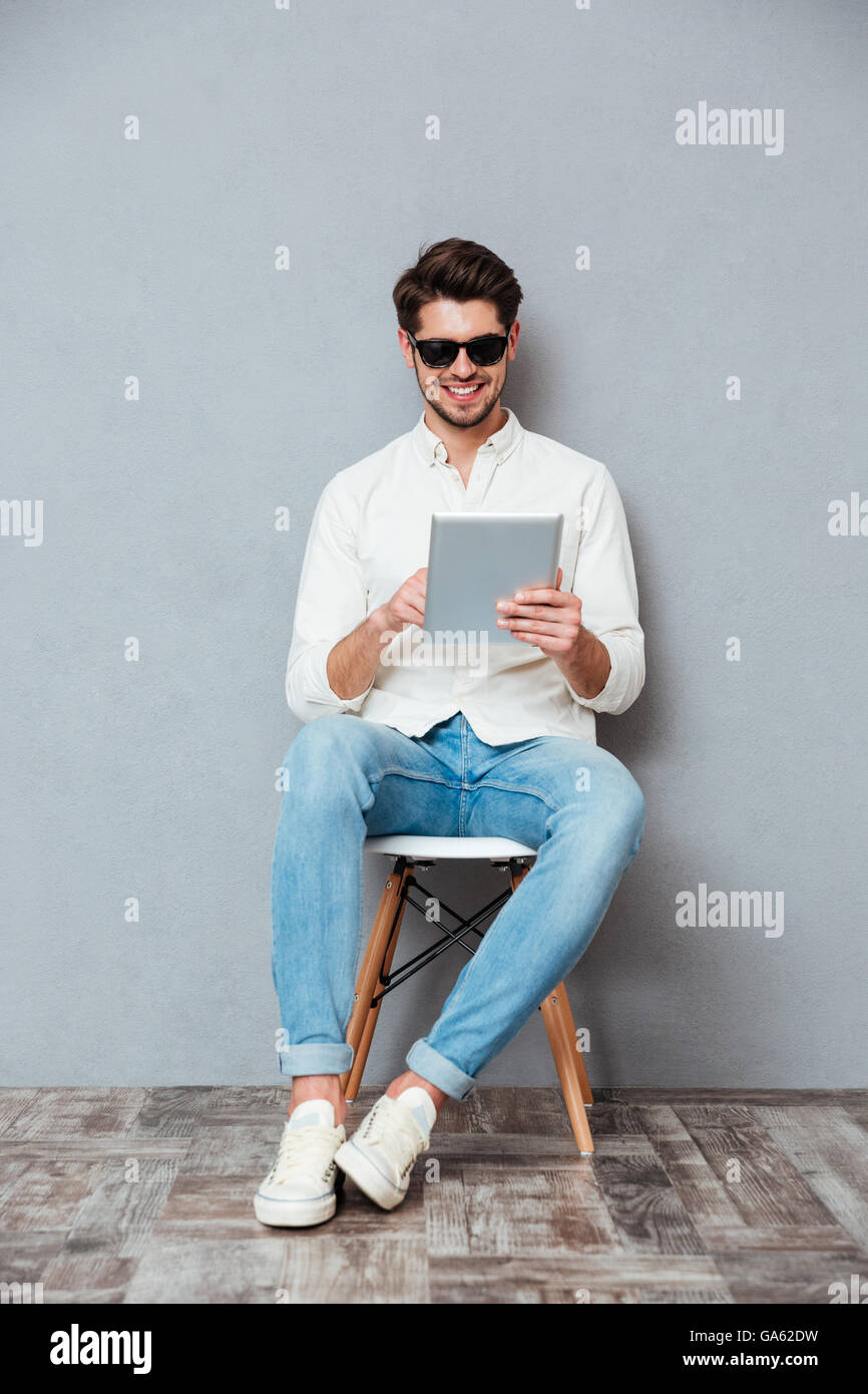 Glücklich Jüngling in Sonnenbrille auf Stuhl sitzend und mit Tablet auf grauem Hintergrund Stockfoto