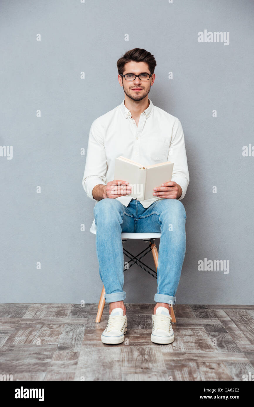 Hübscher junger Mann in Gläsern auf Stuhl sitzen und lesen ein Buch über grauen Hintergrund Stockfoto