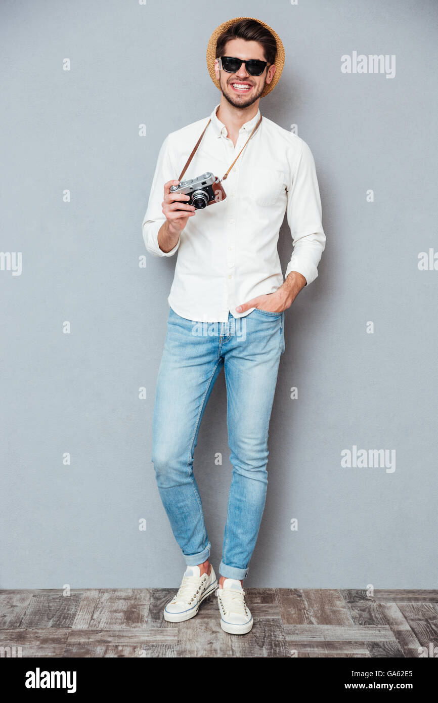 Voller Länge glücklich junger Mann in Hut und eine Sonnenbrille stehen und halten Sie Vintage-Kamera über grauen Hintergrund Stockfoto
