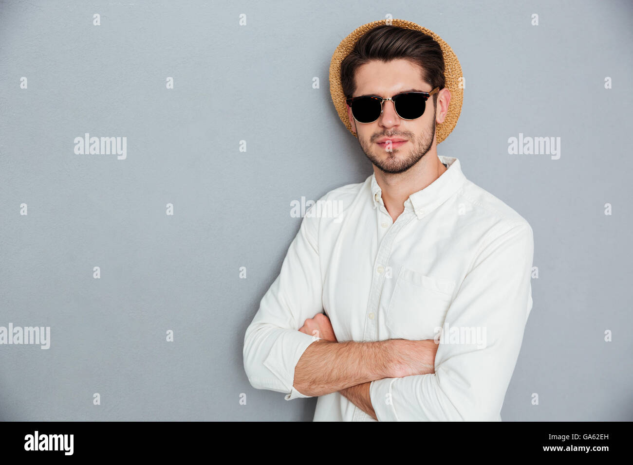 Zuversichtlich, junger Mann in Hut und eine Sonnenbrille mit verschränkten über grauen Hintergrund stehend Stockfoto
