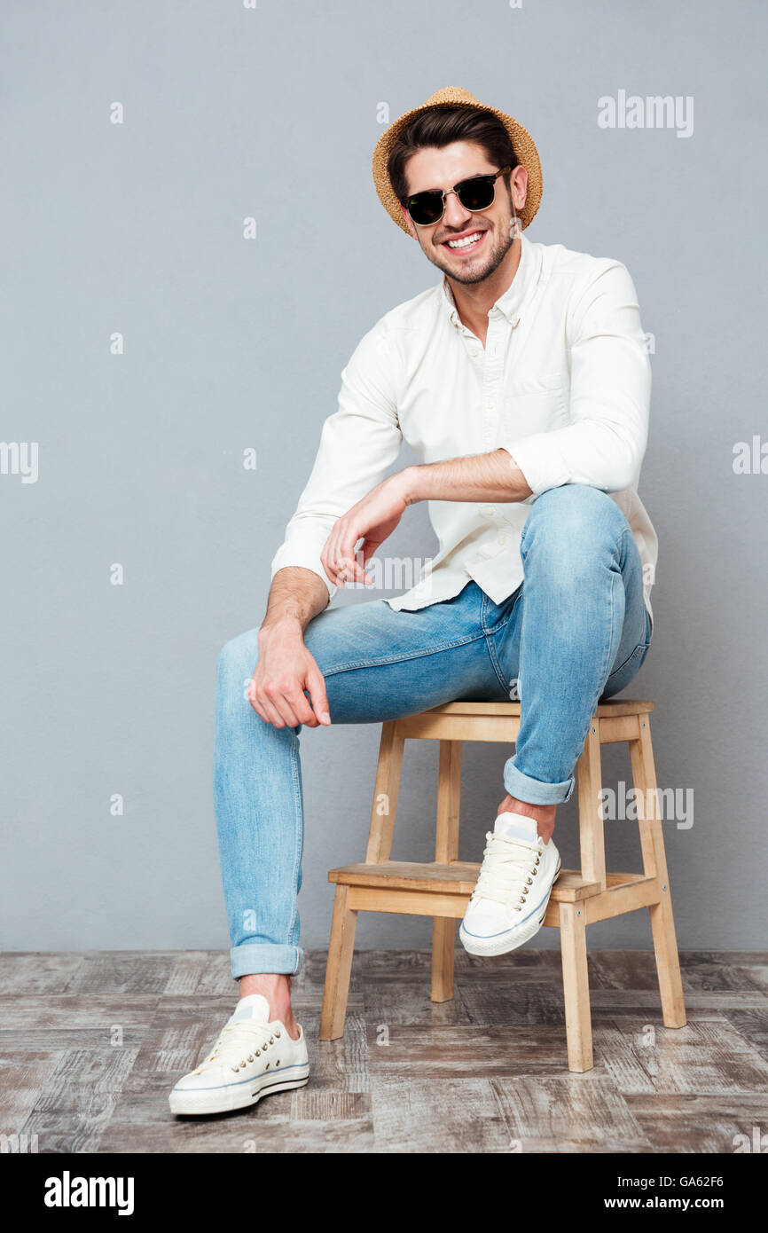 Fröhliche hübscher junger Mann in Hut und Sonnenbrille lächelnd auf grauem Hintergrund Stockfoto
