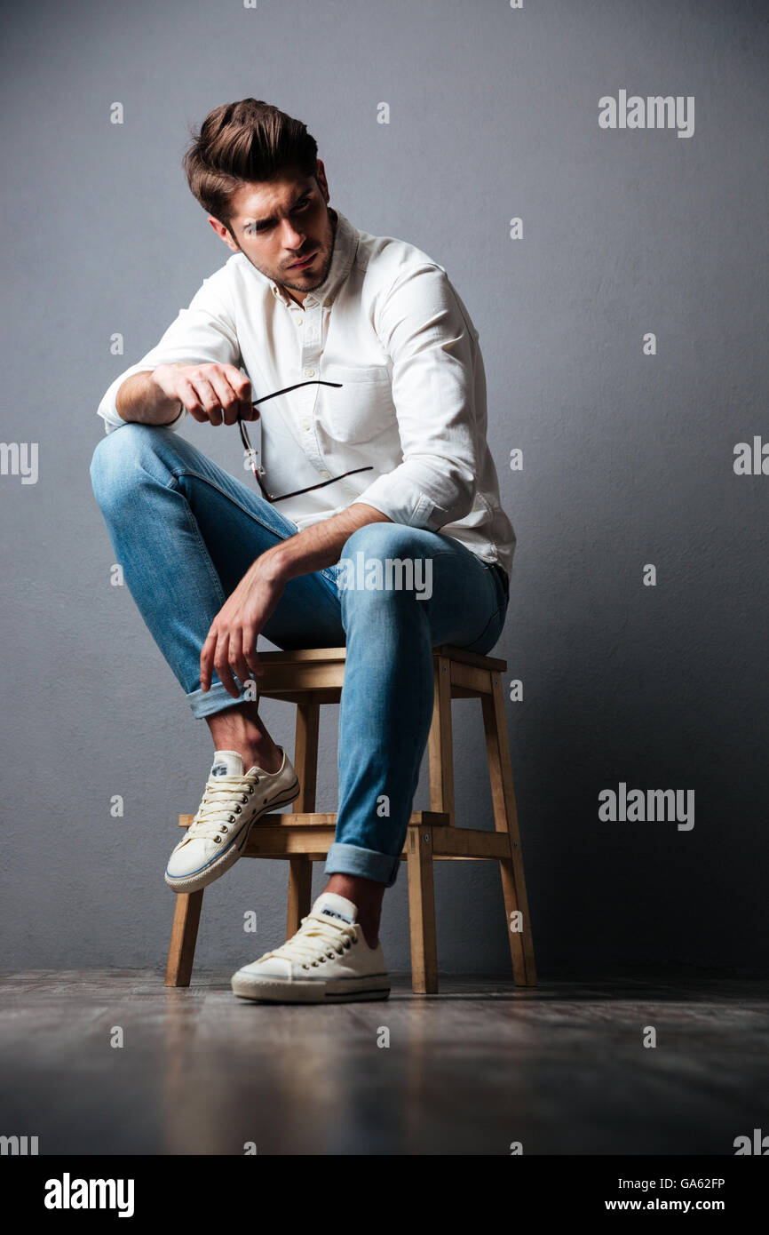 Schwere hübscher junger Mann im weißen Hemd und Jeans sitzen und Sonnenbrillen über grauen Hintergrund zu halten Stockfoto