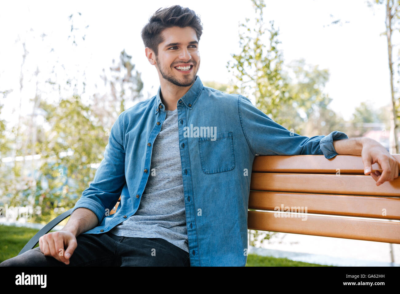 Hübscher junger Mann sitzt auf einer Bank im Park lächelnd Stockfoto