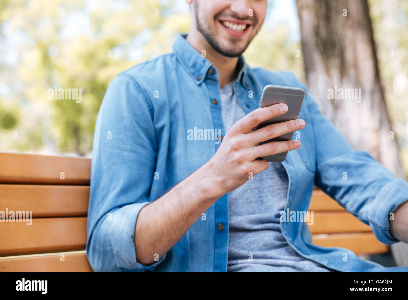 Bild eines lächelnden jungen Mannes mit Smartphone auf der Bank im Freien sitzen abgeschnitten Stockfoto