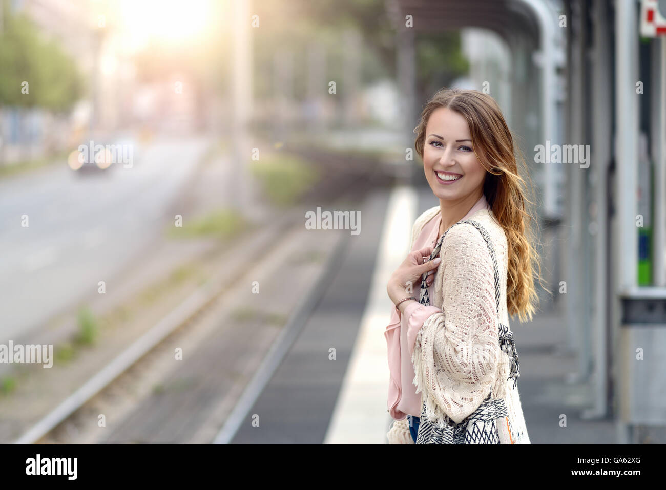 Attraktive stilvolle junge Frau wartet alleine auf einer Plattform an einem kleinen städtischen Bahnhof mit einem glücklichen Lächeln in die Kamera schaut co Stockfoto