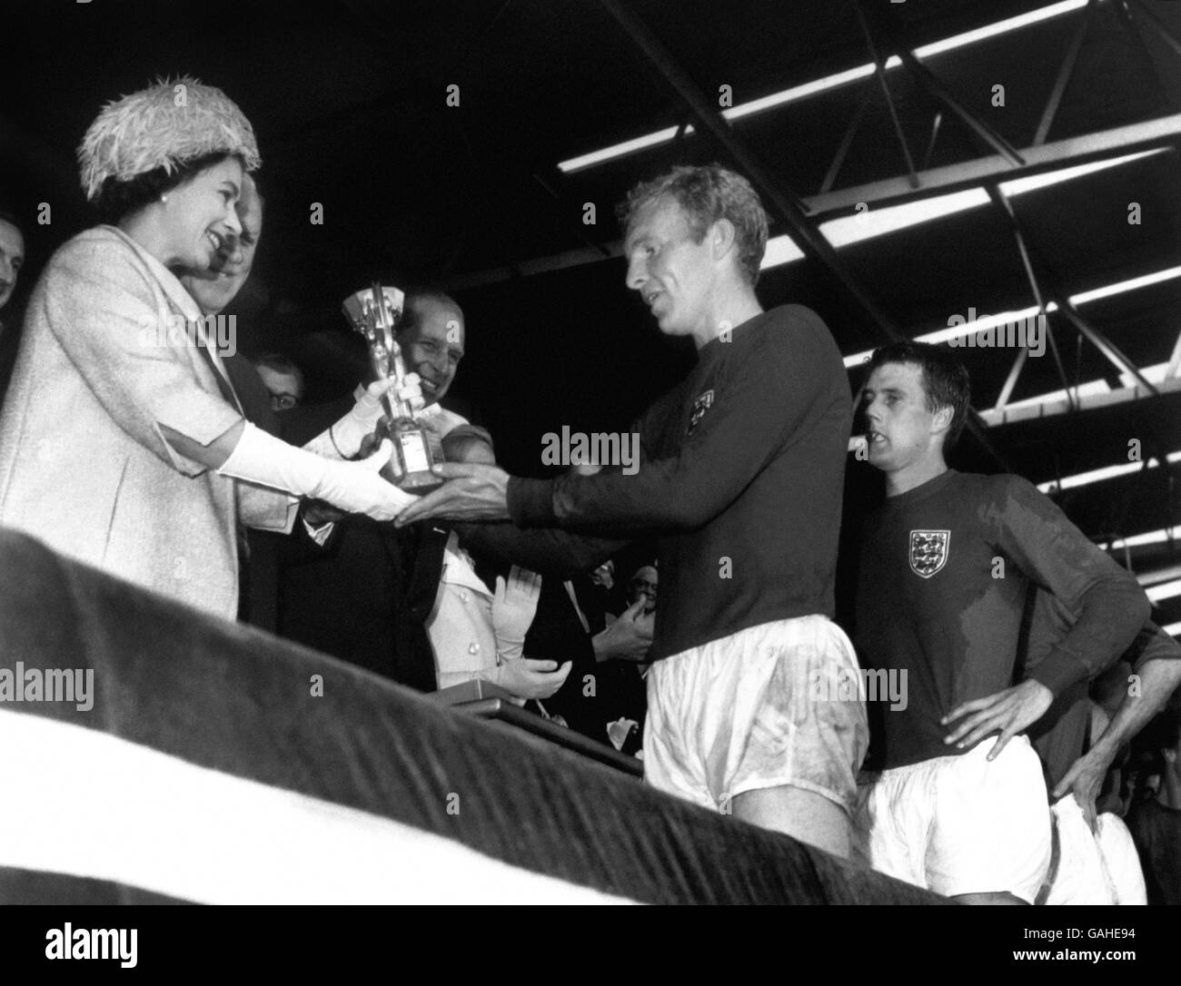 England gegen Westdeutschland - Weltcup-Finale 1966 - Wembley-Stadion. Der englische Kapitän Bobby Moore wird von Ihrer Majestät der Königin mit der Trophäe Jules Rimet überreicht, während Teamkollege Geoff Hurst (r) voller Ehrfurcht daherschaut Stockfoto