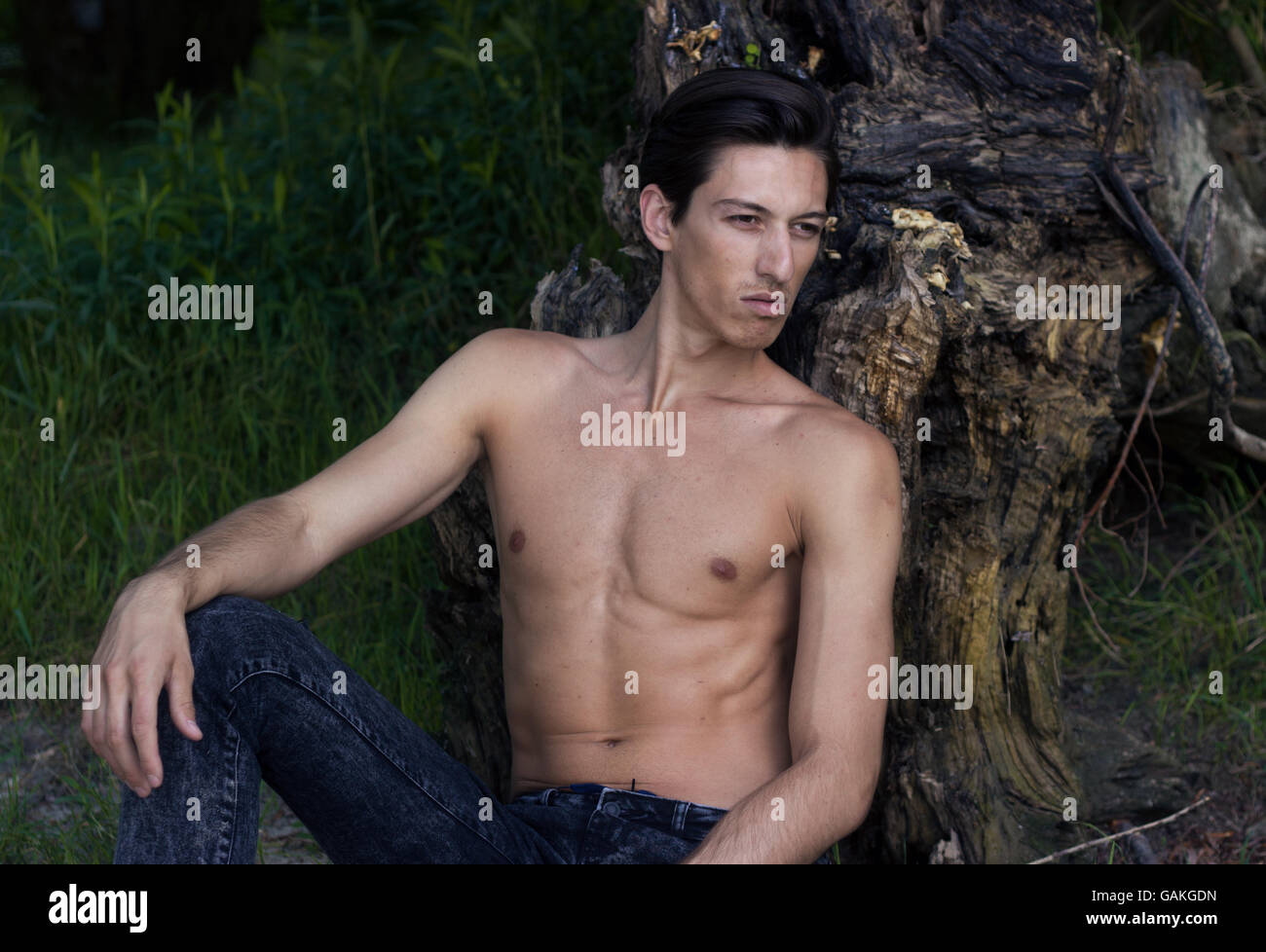 Junger Mann sitzen Sand posiert Skinny slim fit abs Jeans. Kaukasische gebräunte Haut. im Freien, stützte sich auf Baumstumpf sitzend sand Stockfoto