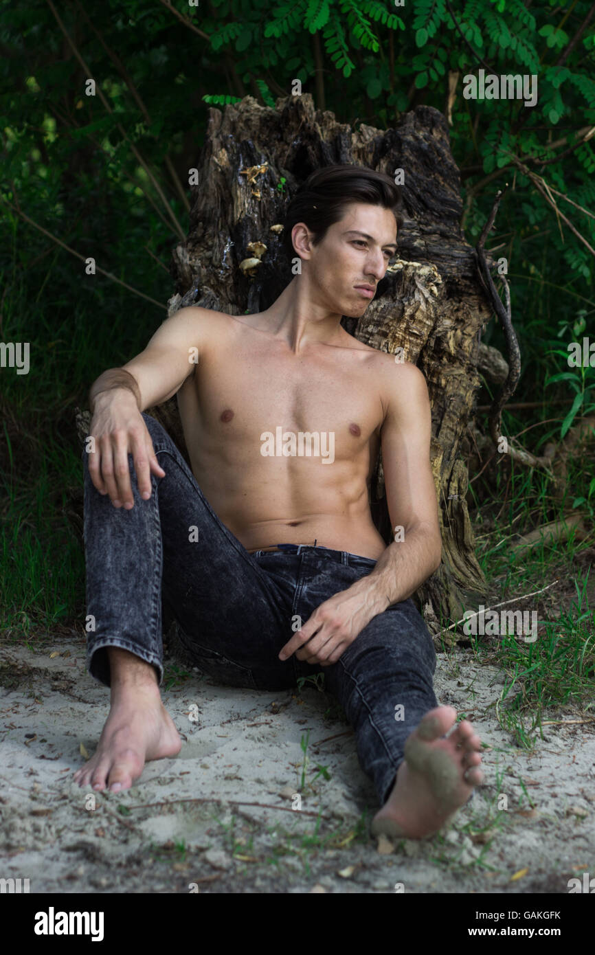 Junger Mann sitzen Sand posiert Skinny slim fit abs Jeans. Kaukasische gebräunte Haut. im Freien, stützte sich auf stumpf, Sand sitzen Stockfoto