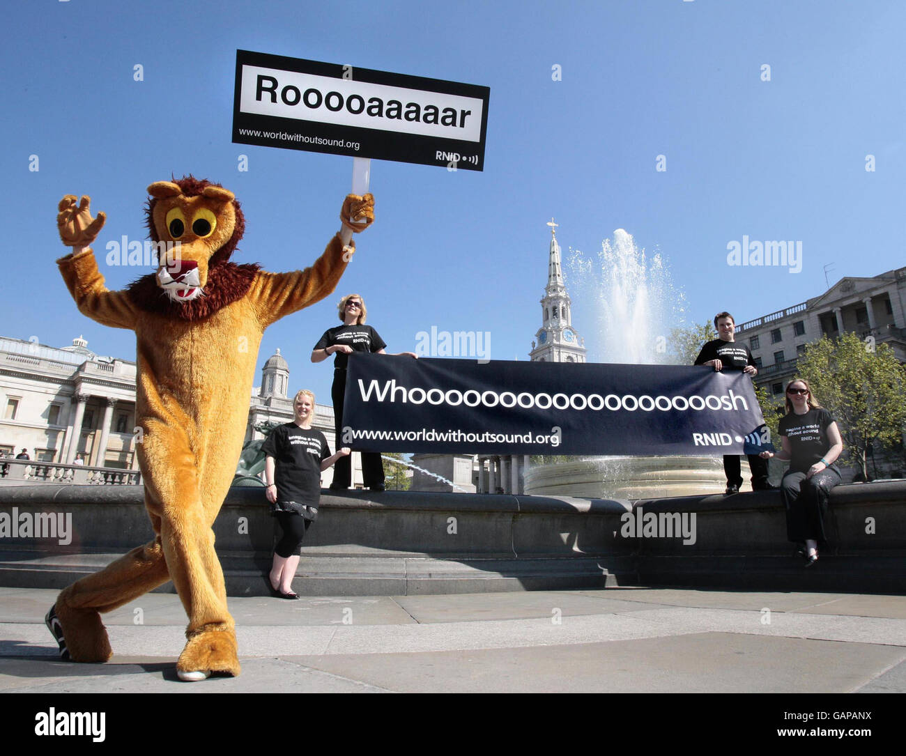 Ein als Löwe gekleideter Aktivist des Royal National Institute for the Deaf hält ein Plakat, auf dem das Brüllen eines Löwen untertitelt wird, während andere Aktivisten ein Transparent halten, das den „Whoosh“ des Brunnens untertitelt, um die „Deaf Awareness Week“ durch die Untertitelung der wichtigsten Wahrzeichen Trafalgar Square, London, hervorzuheben. Stockfoto