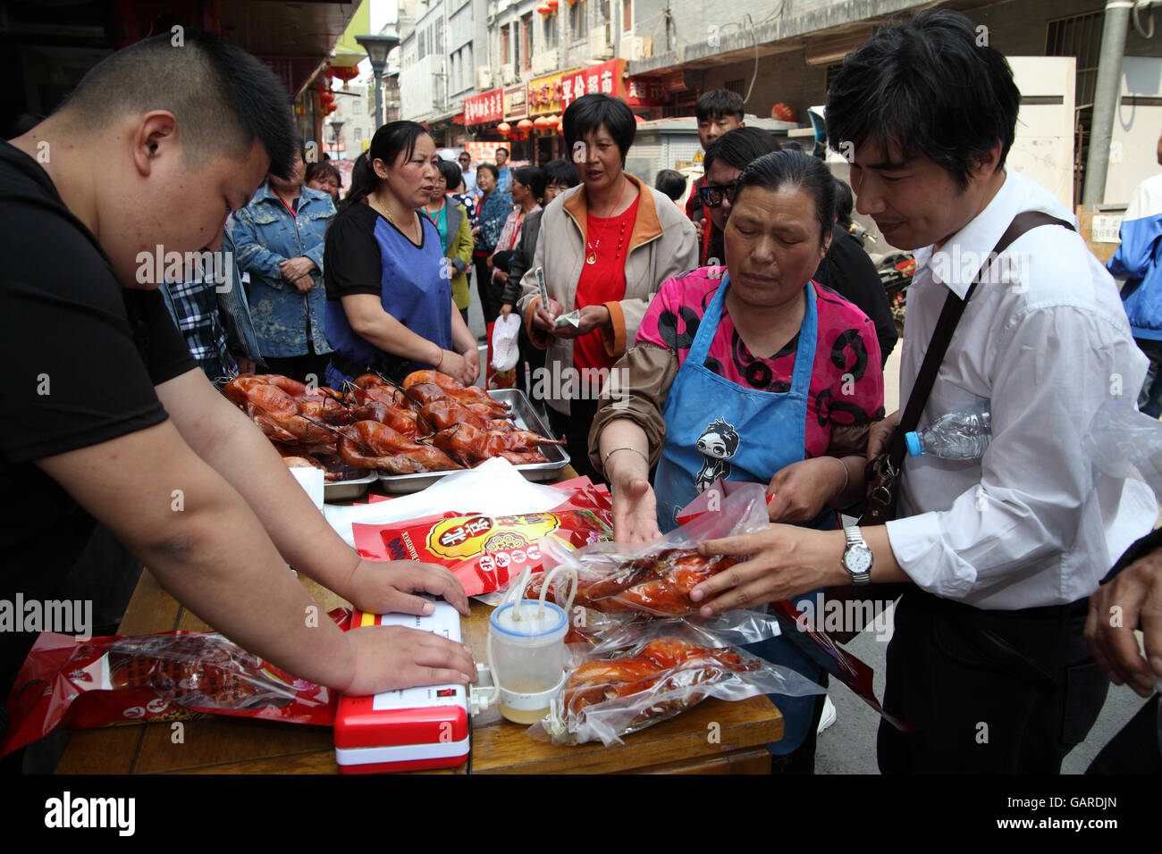 Frau verkauft eingewickelt gegrilltes Hähnchen, ein Mann, ein anderer Mann dichtet eine Plastiktüte mit dem Huhn. Ein Markt unter freiem Himmel, Qibao Old Street, Beijing, China. Stockfoto