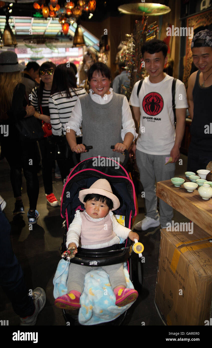 Ein chinesisches Baby sah aus wie eine Puppe sitzt in ihrem Kinderwagen während ihre Eltern über ihr Lächeln in die Kamera.  Der Yuyuan-Viertel, Shanghai, China. Stockfoto