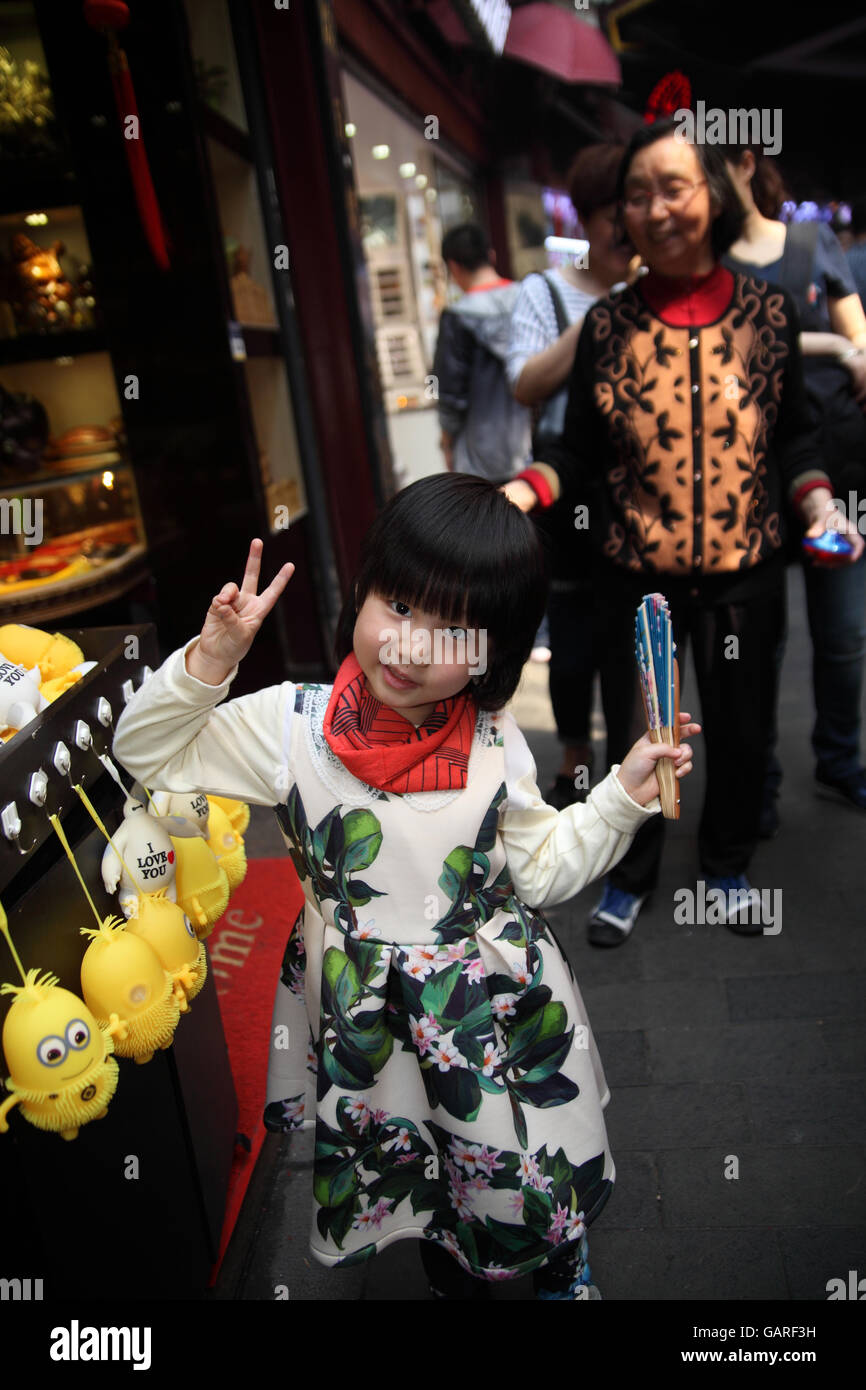 Kleine Mädchen lächelt und macht ein V-Zeichen stehen von Schergen hängen vor einem Geschäft im Quartier Yuyuan, Shanghai, China. Stockfoto