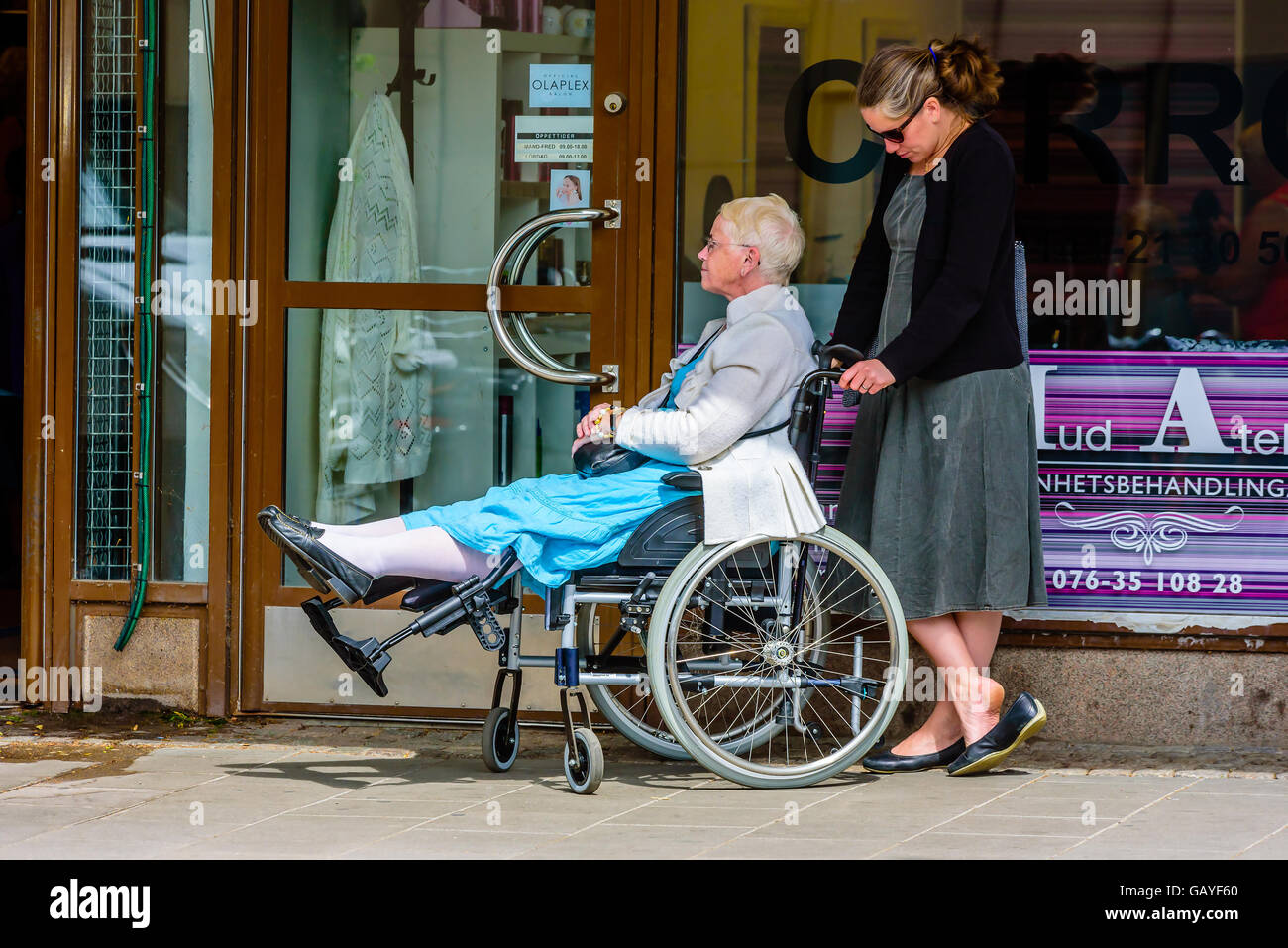Ältere Dame in einem Rollstuhl streckt ihre Beine als sie und ihr Freund oder Helfer wartet Ou Motala, Schweden - 21. Juni 2016: Deaktiviert Stockfoto