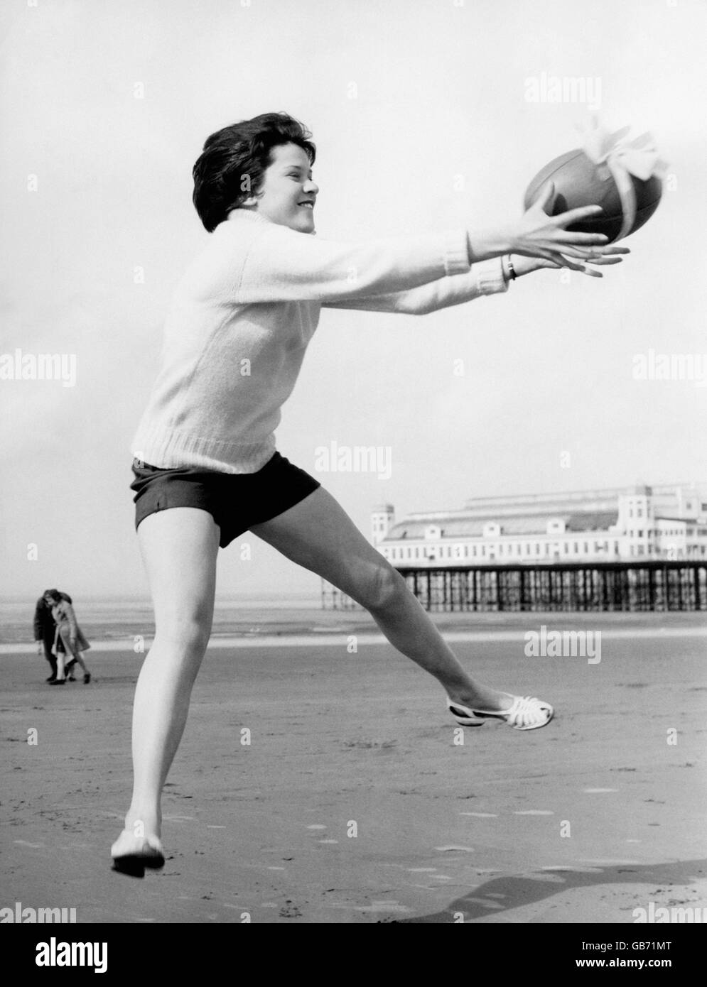 Britische Feiertage - Die Küste - Weston-Super-Mare - 1959. Sally Alford fängt einen Rugby-Ball mit einem dekorativen Band am Strand von Weston-Super-Mare. Stockfoto