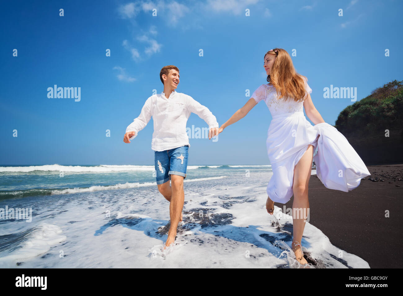 Glückliche Familie Flitterwochen Urlaub - nur verheiratet junger Mann und Frau laufen mit Spaß von schwarzen Sandstrand entlang Meer Surfen. Stockfoto
