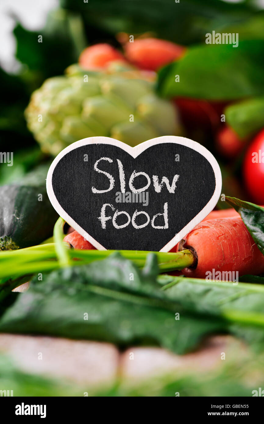 Nahaufnahme von einem herzförmigen Tafel mit dem Text slow Food platziert auf einem Stapel von einigen verschiedenen rohes Gemüse wie Gurken Stockfoto
