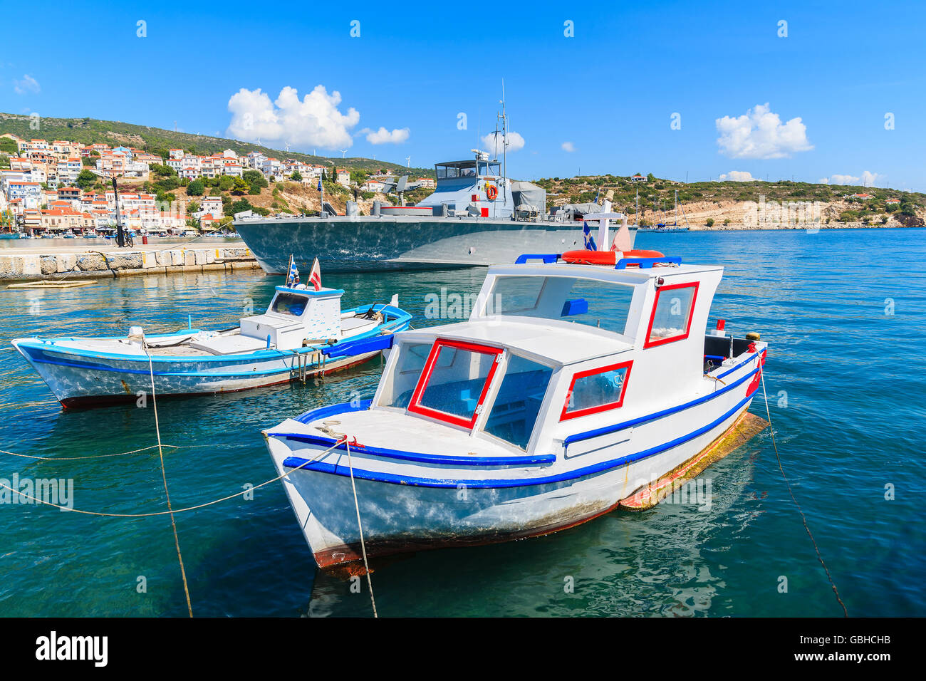Typische bunte Fischerboote mit Kriegsschiff im Hintergrund in Pythagorion Hafen, Insel Samos, Griechenland Stockfoto